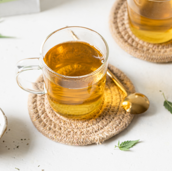 Glas mit aufgebrühtem CBD Bio Hanftee auf geflochtener Unterlage mit goldenem Löffel.