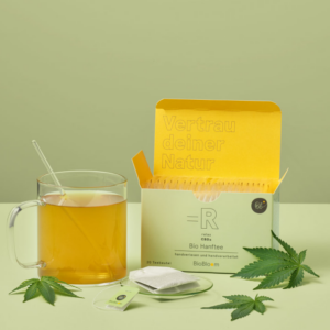 Bio Hanftee relax CBDa von BioBloom im Teeglas serviert, mit Hanfblättern und geöffneter Verpackung