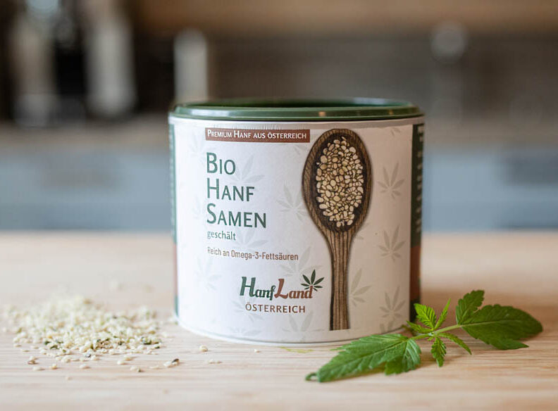 bio-hanfsamen-geschaelt-oesterreich-hanfland.jpg Bio Hanfsamen geschält von Hanfland in der Dose, daneben lose Samen und ein grünes Hanfblatt auf hellem Holzgrund – direkt aus Österreich.