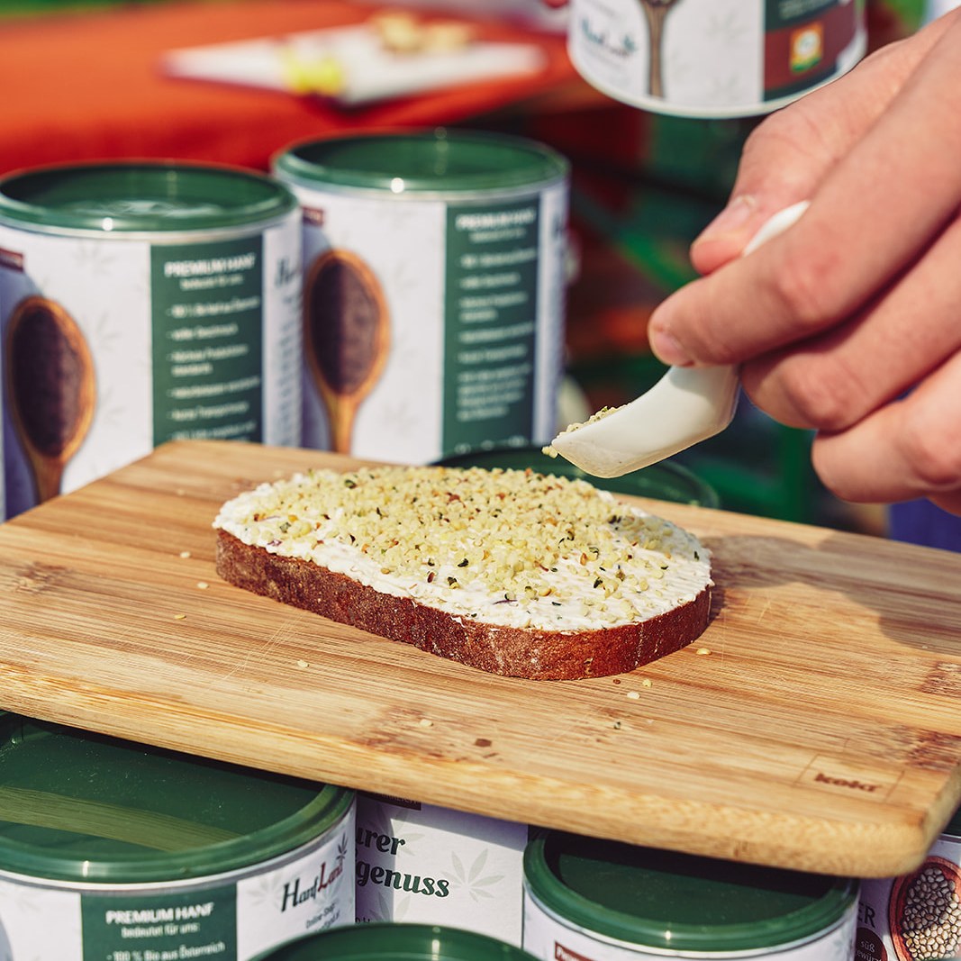 Brotscheibe mit Frischkäse wird mit geschälten Bio Hanfsamen bestreut, Hanfland Dosen im Hintergrund.